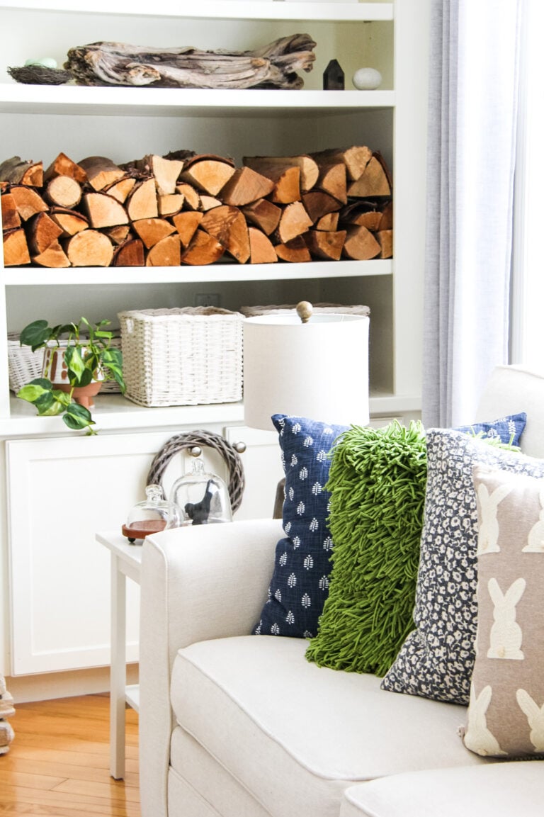 blue, tan and green cushions sit on a white couch. in the background is a white shelf with stacks of wood, a green plant and white picture frames