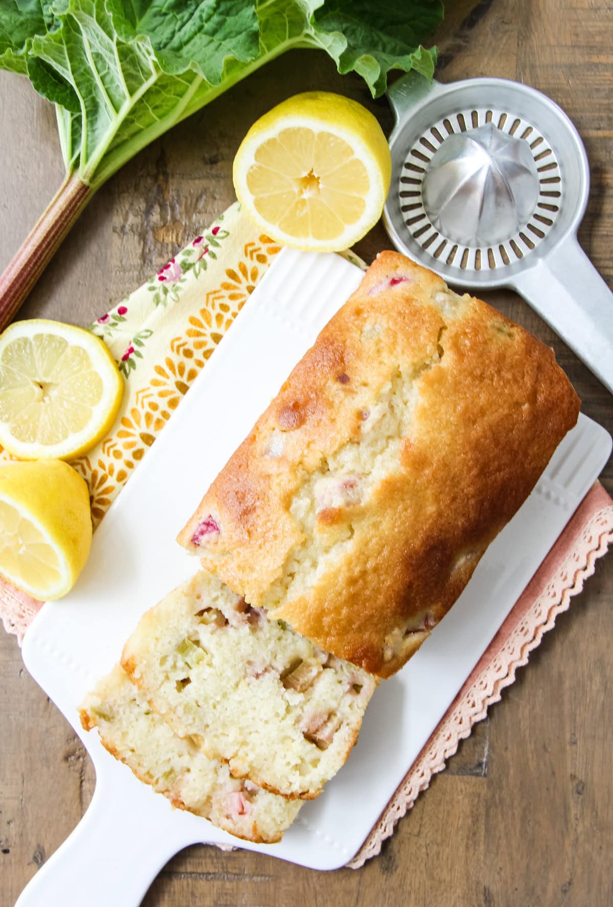 Rhubarb Lemon Bread cut into slices, sitting on a white cutting board next to fresh lemons and rhubarb