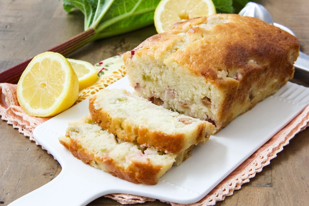 Rhubarb Lemon Bread cut into slices, sitting on a white cutting board next to fresh lemons and rhubarb